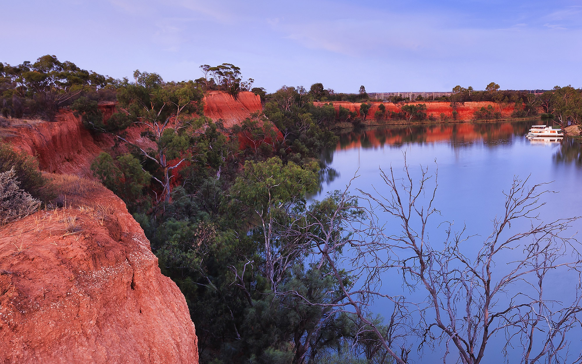 Red cliffs of Murray river on the border between Victoria and New South Wales states of Australia. Elevated red bank of the river overlooks river bend with cruise ship at sunset.